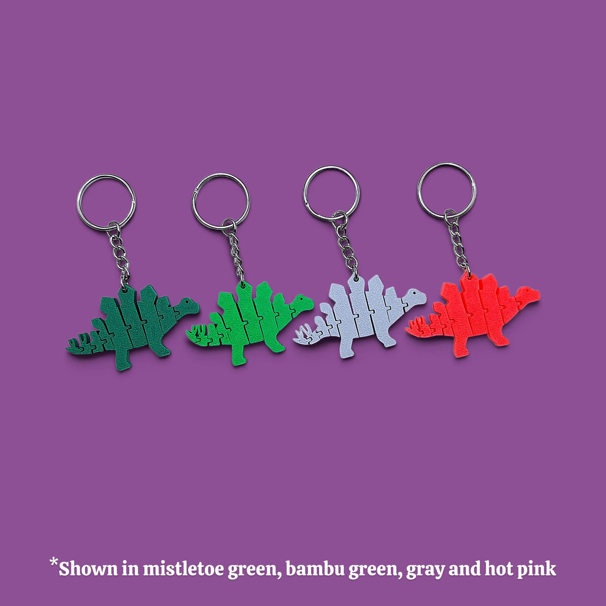 Four dinosaur-shaped keychains in different colors (mistletoe green, bamboo green, gray, and hot pink) arranged in a row on a solid purple background, with a text overlay at the bottom stating '*Shown in mistletoe green, bambu green, gray and hot pink'.
